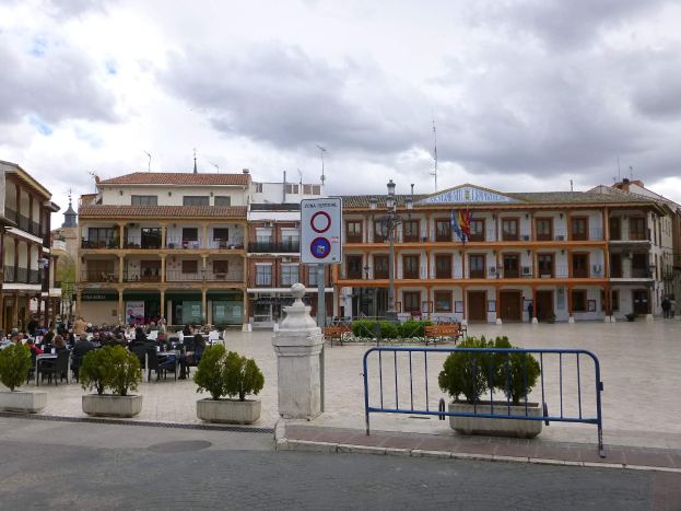 Ein belebter Stadtplatz mit Menschen auf Stühlen sitzend und stehend, umgeben von Topfpflanzen, Metallabsperrungen, Schildern, Straßenmasten mit Fahnen, Gebäuden und einem bewölkten Himmel.