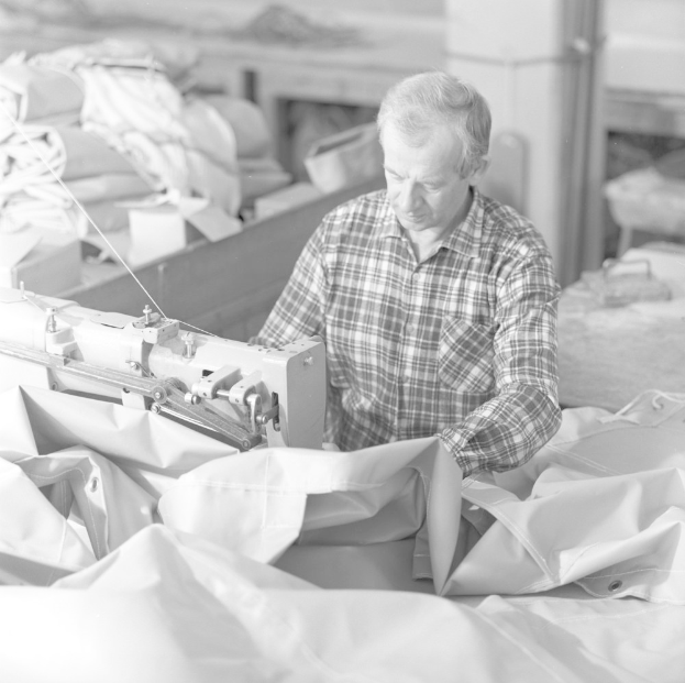 Ein Mann bedient eine Nähmaschine in einer Fabrik, umgeben von Kleidung und anderen Gegenständen, dargestellt in Schwarz-Weiß.