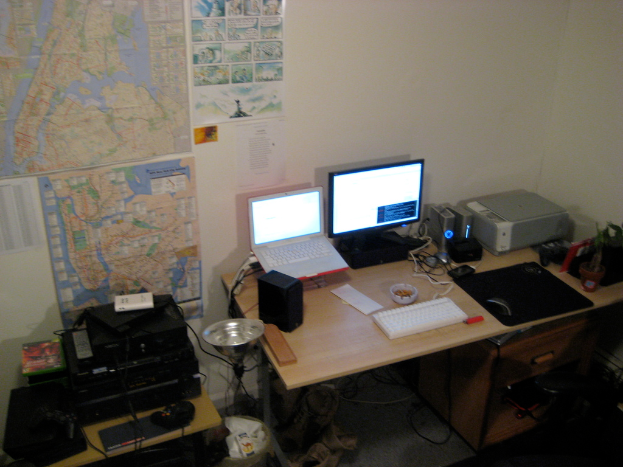 Ein Raum mit einem Tisch, auf dem ein Laptop, Lautsprecher, Monitor, Tastatur, Ball, Maus, Scanner und Papiere stehen; ein weiterer Tisch mit Elektronikgeraten links unten; und eine Wand mit Charts und Postern im Hintergrund.