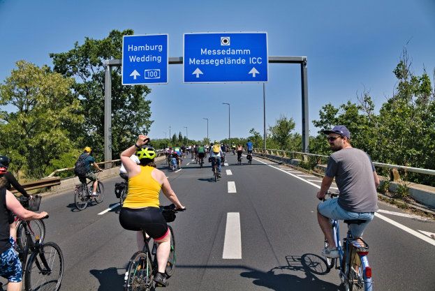 Eine Gruppe von Radfahrern mit Helmen fährt auf einer Straße mit einer Begrenzung auf einer Seite und Bäumen auf der anderen unter einem klaren blauen Himmel mit Laternen und einem Schild, das eine Radtour in Hamburg anzeigt.