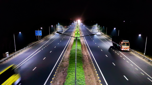 Ein Lastwagen fährt nachts auf einer dunklen Autobahn, beleuchtet von Straßenlaternen, umgeben von Gras, Pfählen und Schildern.
