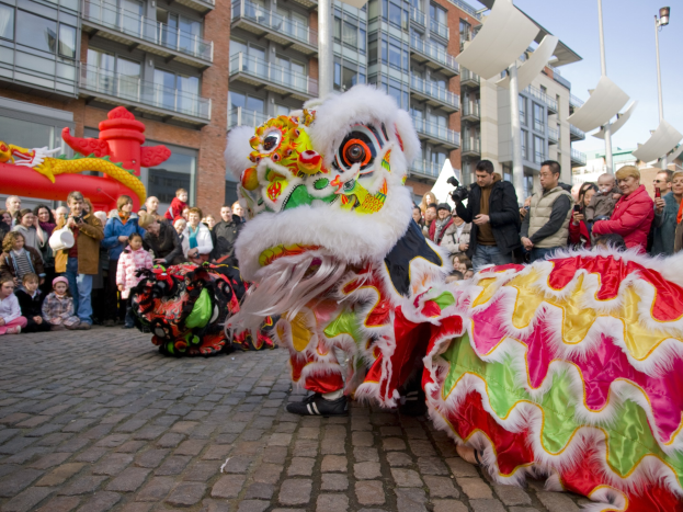 Ein lebendiges chinesisches Neujahrsfest in Amsterdam mit einem Löwen tanzen im Vordergrund und einer Menge Menschen drumherum, einige halten Kameras, vor einem Hintergrund aus Gebäuden, Laternenmasten und einem klaren blauen Himmel.