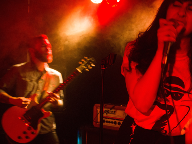 Eine Frau singt in ein Mikrofon auf einer Bühne, wöhrend ein Mann im Hintergrund eine Akustikgitarre spielt, mit Deckenlampen, die die Szenerie beleuchten.