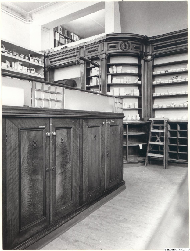 Schwarzes und weißes Foto eines alten Apothekeninneren mit einer Theke, Schränken, einer Leiter und Regalen, die mit verschiedenen Gegenständen gefüllt sind, mit Text oben.