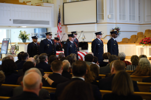 Gruppe uniformierter Personen vor sitzenden Trauergästen bei einer Trauerfeier, mit Blumenarrangements, einer Fahne, einem Bildschirm und einer Wand im Hintergrund.