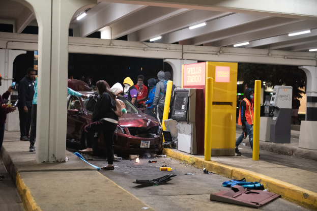 Eine Gruppe von Menschen steht um ein beschädigtes Auto herum, das in eine Tankstelle gekracht ist. Es gibt verstreute Gegenstände auf dem Boden und Säulen, Maschinen und Deckenlampen im Hintergrund.