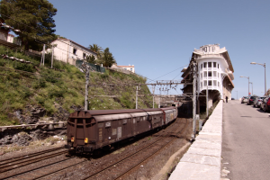 Güterzug fährt auf Gleisen neben einem Gebäude mit parkenden Autos auf einem Bahnsteig auf der rechten Seite, vor einer Kulisse aus städtischen Gebäuden, Bäumen, Strommasten und einem klaren Himmel.