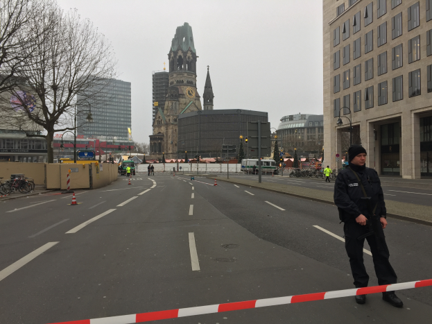 Ein Polizist in schwarzer Uniform und Mütze steht auf einer Straße in Berlin umgeben von Verkehrskegeln, Fahrrädern, Laternenpfählen, Bäumen und Gebäuden, mit einem Uhrenturm und einem klaren blauen Himmel im Hintergrund.