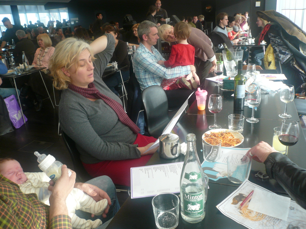 Ein Innenblick in ein belebtes Restaurant mit Menschen an Tischen, die mit Gläsern, Tellern mit Essen, Getränkeflaschen, Speisekarten und anderen Gegenständen gedeckt sind.