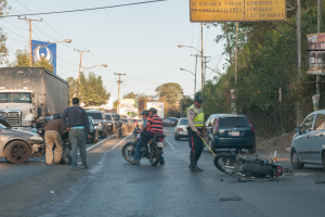 Gruppe von Menschen um ein verunglücktes Motorrad am Straßenrand mit mehreren Fahrzeugen, darunter ein Lastwagen, im Hintergrund und Bäumen, Masten, Lichter, Schilder und Himmel.