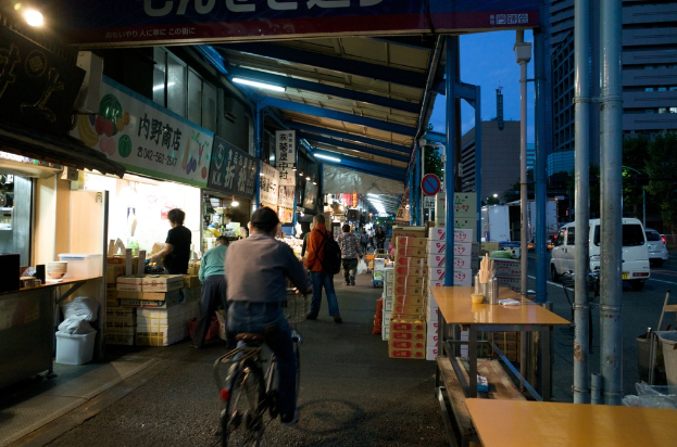 Ein Gehweg mit Geschäften auf der linken Seite und Kisten, einem Tisch, einer Straße mit Autos, Bäumen und einem Gebäude auf der rechten Seite; eine Person auf einem Fahrrad und eine weitere Person in einem orangen Hemd und Jeans mit einer Tasche sind zu sehen.