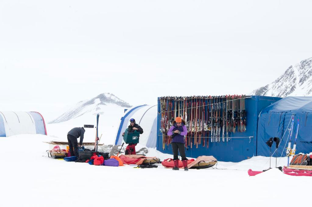 Drei Personen stehen auf einer verschneiten Landschaft mit Taschen, die herumliegen, im Hintergrund Zelte mit Skiern darauf und schneebedeckte Hügel unter einem klaren Himmel.