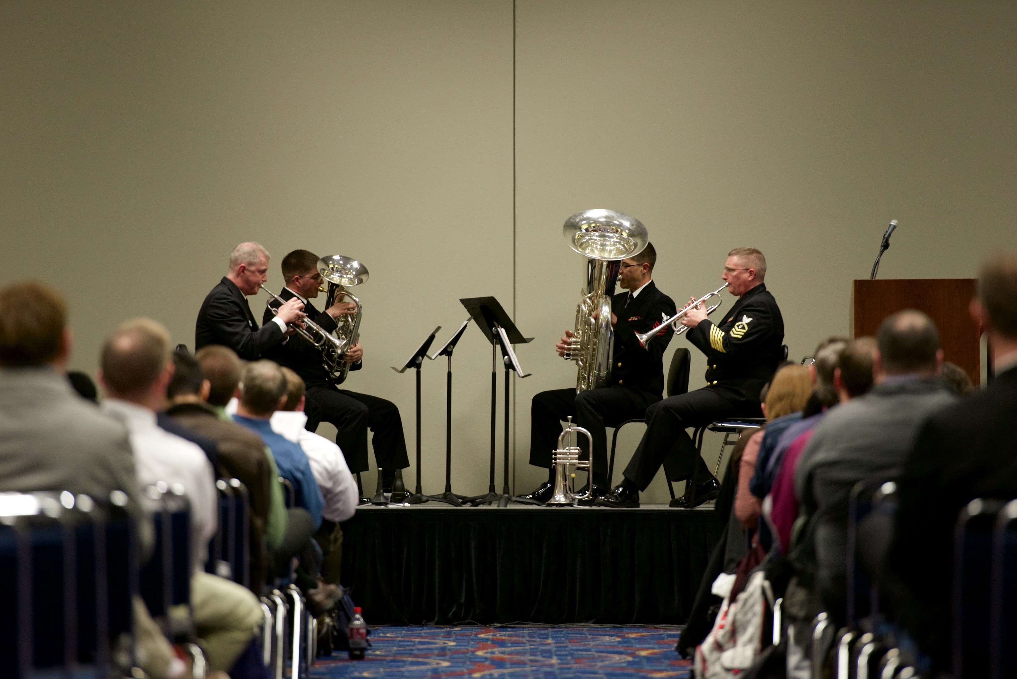 Eine Gruppe von Männern, die auf einer Bühne Blasinstrumente spielen, mit Notenständern, einem Podium mit Mikrofon und einem Publikum, das in Stühlen sitzt und einer Wand im Hintergrund.