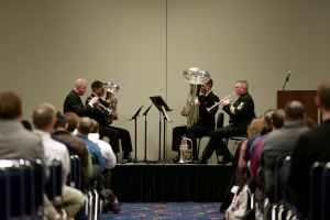 Eine Gruppe von Männern, die auf einer Bühne Blasinstrumente spielen, mit Notenständern, einem Podium mit Mikrofon und einem Publikum, das in Stühlen sitzt und einer Wand im Hintergrund.