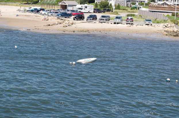 Ein Boot schwimmt auf dem Meer mit parkenden Autos, Bäumen, Pflanzen und Gebäuden im Hintergrund.