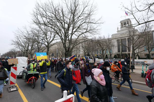 Eine große Gruppe von Menschen marschiert auf einer Demonstration durch eine Straße in Washington, D.C., mit Schildern, Bannern und Fahrrädern, vor einem Gebäude, Bäumen, Schildern und einem klaren blauen Himmel.