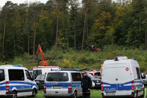 Eine Gruppe von Polizeiwagen in einer Reihe auf einem Parkplatz mit einer Person im Vordergrund, Bäumen, Pflanzen, einem Bagger und einem klaren Himmel im Hintergrund.