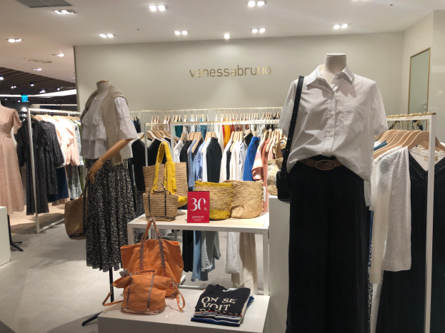 Ein Vanessa Bruno Geschäft in Singapur mit Schaufensterpuppen in Kleidern, hängenden Kleidern, Taschen auf Tischen, einer Texttafel an der Wand, einem Spiegel auf der rechten Seite und Deckenleuchten.