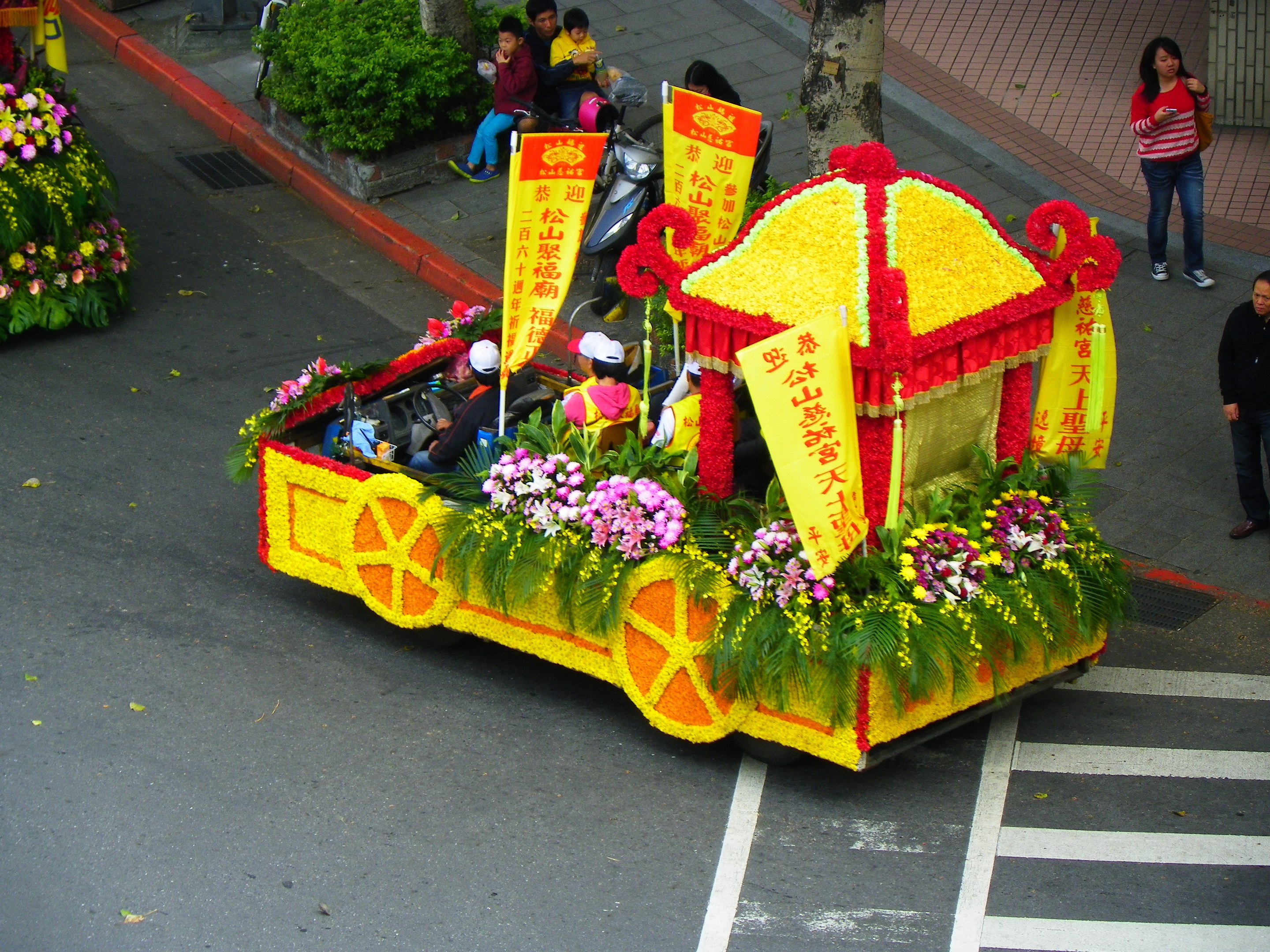 Ein Paradewagen mit Blumen und Bannern geschmückt, auf dem Reiter sitzen, mit Zuschauern rechts und Grün im Hintergrund.