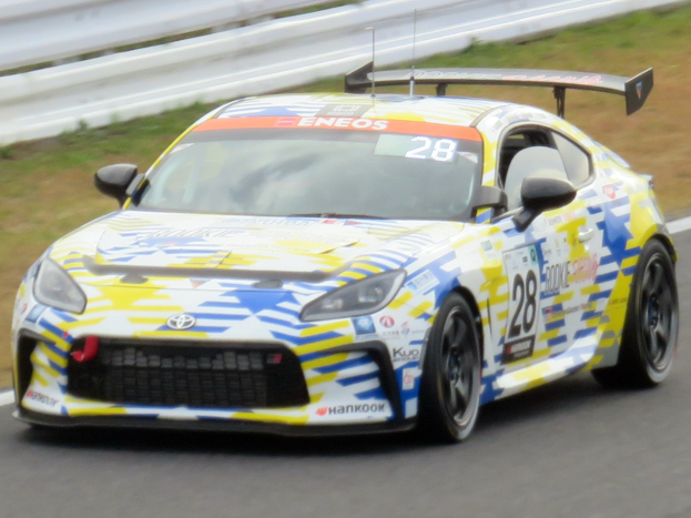 Ein weißer Toyota Supra GT3-Rennwagen mit Sponsor-Text und Nummern auf einer Rennstrecke, umgeben von einer Begrenzung und Gras.