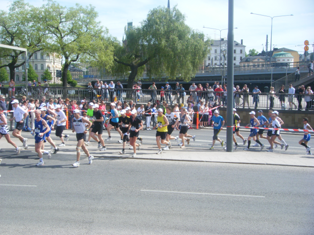 Eine Gruppe von Läufern bei einem Marathon auf einer Straße, umgeben von einem Metallzaun, einem Tor, einer Schleife an einem Pfahl, Zuschauern auf dem Gehweg, einer Metallabsperrung, Pfählen, Straßenlaternen, Schildern, einer Brücke, Gebäuden mit Fenstern, Bäumen und einem bewölkten Himmel.