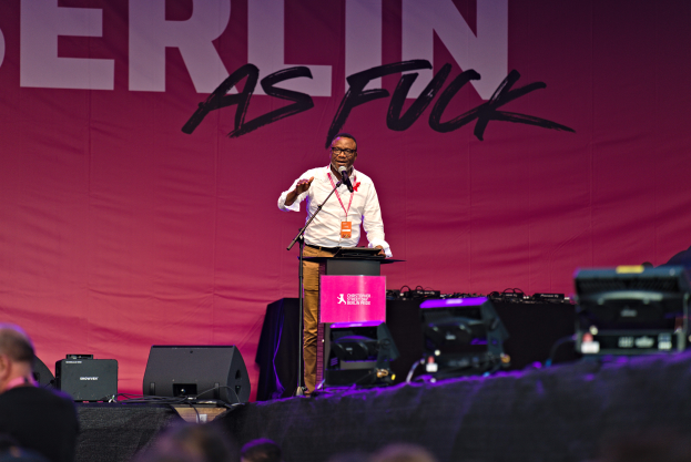 Ein Mann in weißem Hemd und braunen Hosen steht auf einer Bühne und hält ein Mikrofon in der Hand, hinter ihm eine pinkfarbene Fahne mit der Aufschrift "Berlin as Fuck" und Lautsprecher auf der Bühne.
