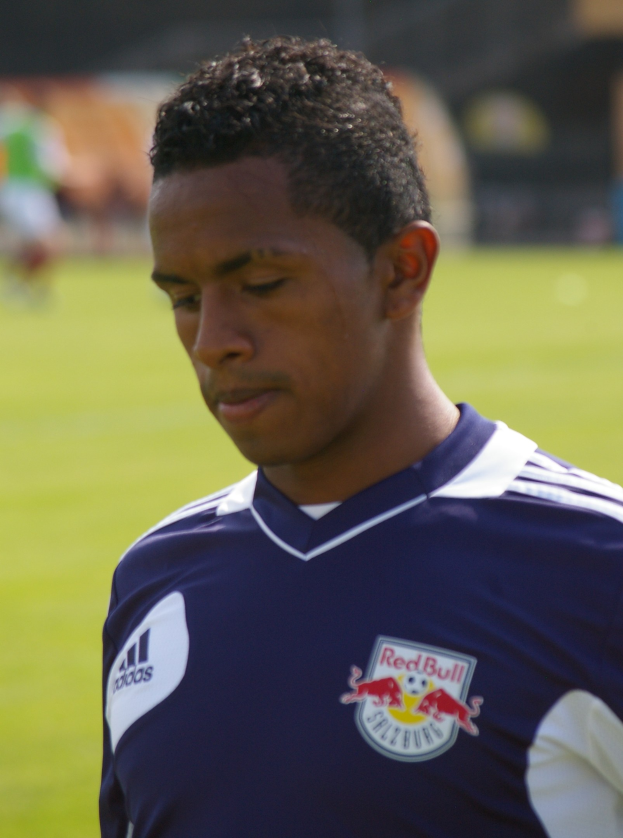 Junger Mann in blauem T-Shirt auf einem Fuballfeld mit entschlossener Miene, unscharfer Hintergrund.