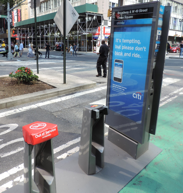 Eine belebte Stadtstraße mit Fußgängern auf dem Gehweg, Fahrzeugen auf der Straße und einem Bushaltestellen-Schild mit der Aufschrift "Citi" neben einem Müllcontainer.