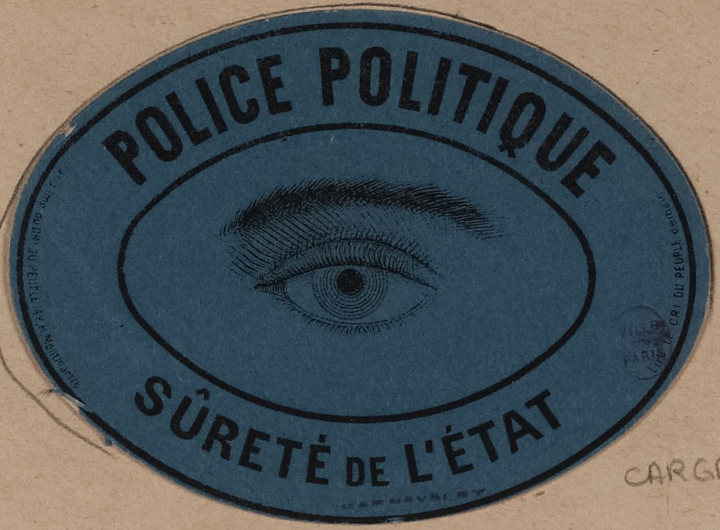 Braune Kartonbox mit blauem Aufkleber mit der Aufschrift "Polizei Politique Sureté de L'Etat" und einem Auge-Symbol.
