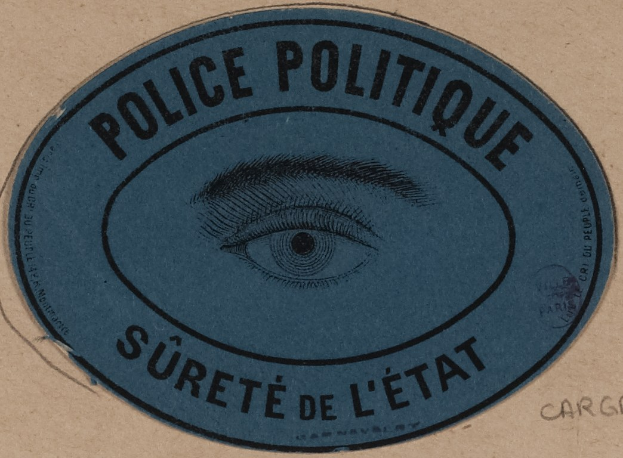 Braune Kartonbox mit blauem Aufkleber mit der Aufschrift "Polizei Politique Sureté de L'Etat" und einem Auge-Symbol.