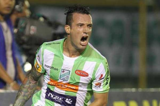 Fussballspieler in grün-weissem Trikot mit Tattoos auf dem Arm, der einen Torerfolg feiert, mit unscharfem Hintergrund mit Menschen und einer Tafel.