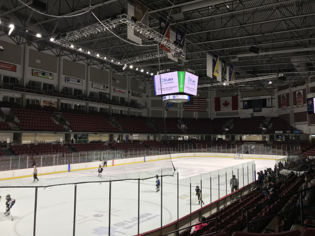 Eishockeyspiel in einer großen Arena mit einem zentralen Tor, Zuschauern auf Stühlen, Deckenbeleuchtung und Trägern sowie Wandbannern mit der Aufschrift "St. Louis Blues Arena."