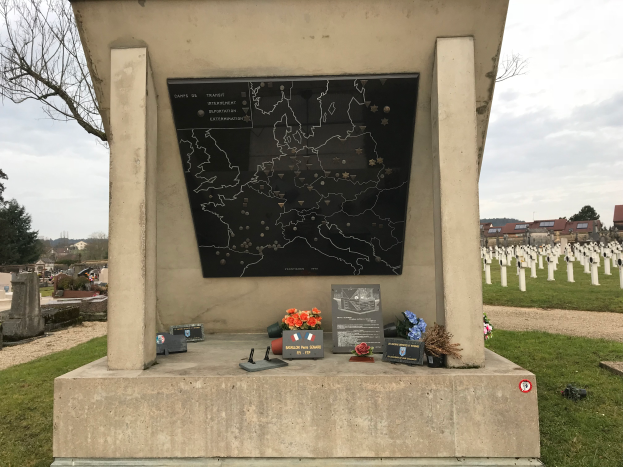 Ein Denkmal auf einem Friedhof mit einer Karte darauf, umgeben von Blumensträußen und anderen Gegenständen, mit Gräbern, Bäumen, Gebäuden und einem klaren blauen Himmel im Hintergrund, das das Grab eines deutschen Soldaten markiert, der im Zweiten Weltkrieg getötet wurde.