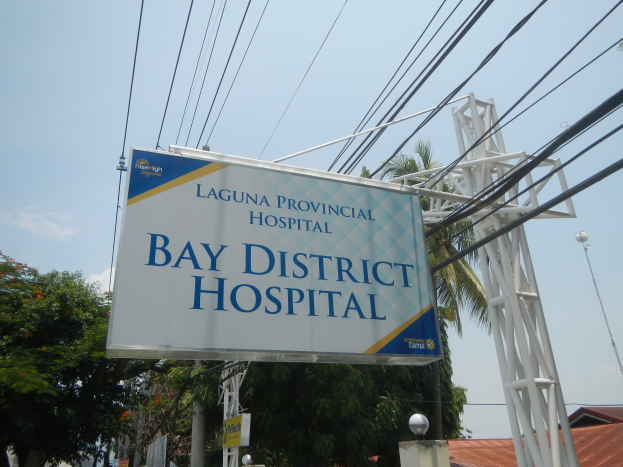 Krankenhaus im Landkreis mit einem Schild, Strommasten, Laternen, Bäumen, Gebäuden und einem bewölkten Himmel im Hintergrund.