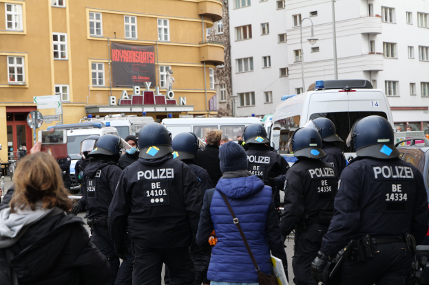 Eine Gruppe von Polizisten steht vor einer Menge von Menschen, die Helme und Jacken tragen, während einer Demonstration in Berlin, Deutschland, mit Fahrzeugen, Gebäuden, Laternenmasten und einer Fahne mit Text im Hintergrund und einer Person mit einer Kamera.