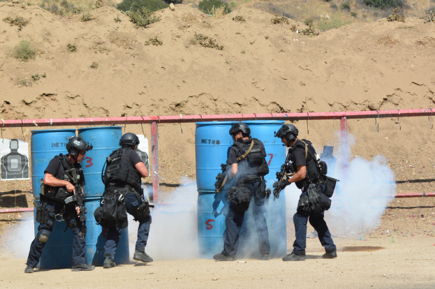 Eine Gruppe von Polizisten in schwarzer Einsatzkleidung, jeder mit einer Waffe in der Hand, steht zusammen auf einem Feld mit blauen Tonnen, einem roten Zaun, einem Plakat, Pflanzen und einem Berg im Hintergrund.