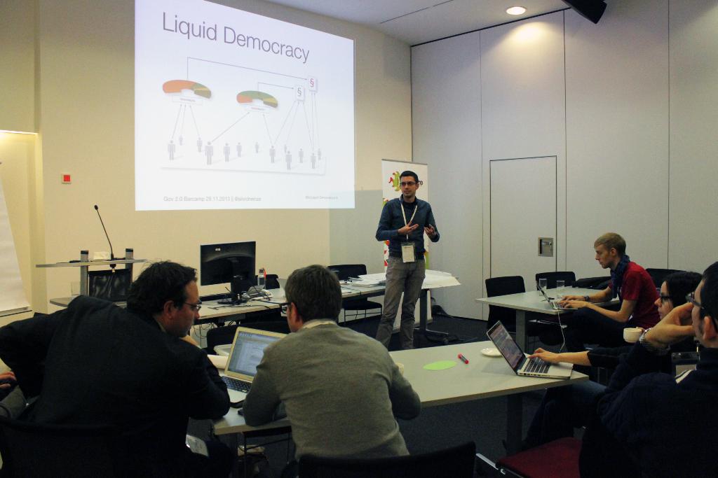 Fünf Personen in einem Konferenzsaal, die einem stehenden Redner zuhören, mit Tischen, die Laptops, Marker, Papiere, einen Monitor, ein Mikrofon und eine Projektionswand mit Text enthalten.