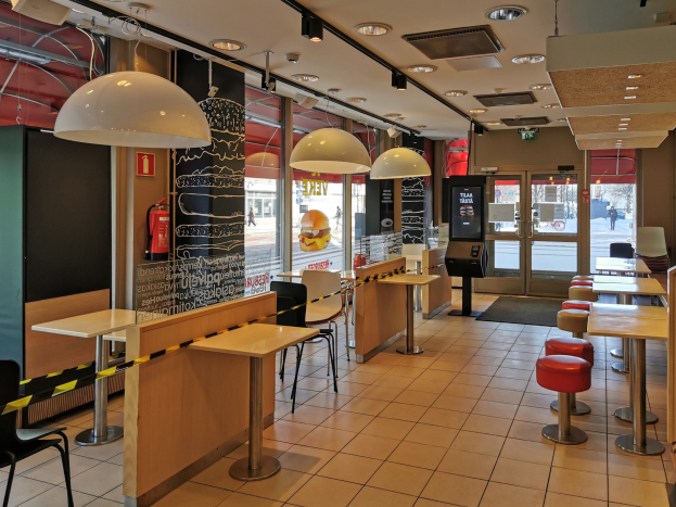 Innenraum eines Fast-Food-Restaurants mit Tischen, Stühlen, Hockern, einem Boden, Deckenleuchten, einem wandmontierten Feuerlöscher, Glastüren und zusätzlichen Gegenständen.