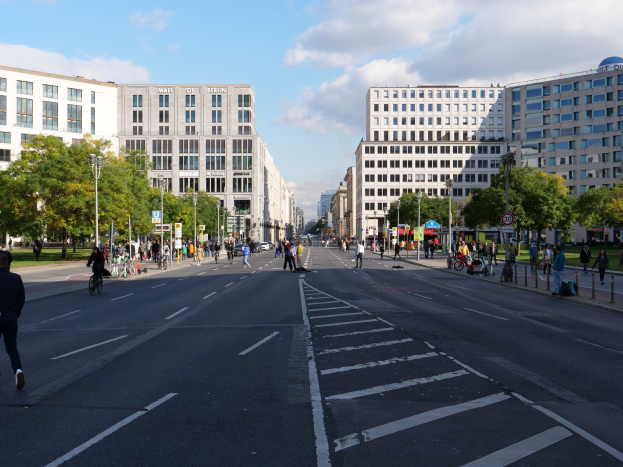 Eine belebte Stadtstraße in Berlin, Deutschland, mit Fußgängern und Radfahrern auf der Straße, hohen Gebäuden, Bäumen, Laternenmasten und Schildern unter einem bewölkten Himmel.