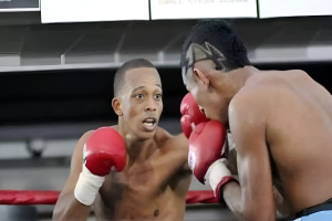 Zwei Boxer, die in einem Boxring kämpfen, mit einer Zuschauermenge und einer Wand mit Text und Logos im Hintergrund.