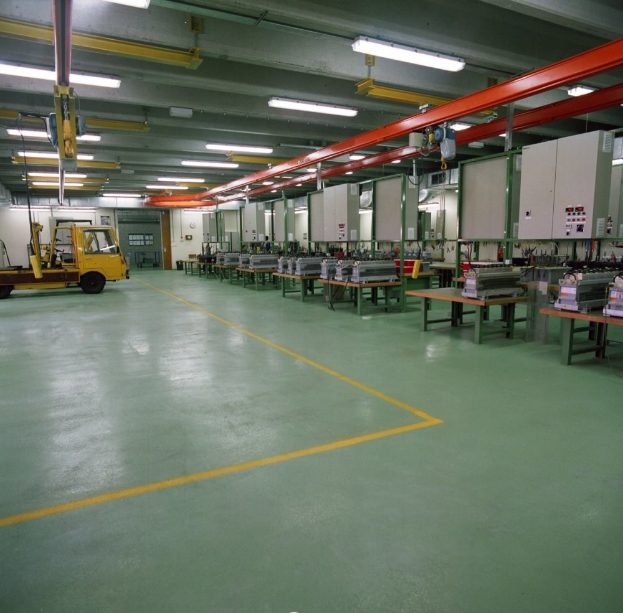 Ein großes Fabrikinnere mit einem Gabelstapler in der Mitte, Tische mit Gegenständen, Maschinen und Deckenleuchten, das zeigt den Boden unten.