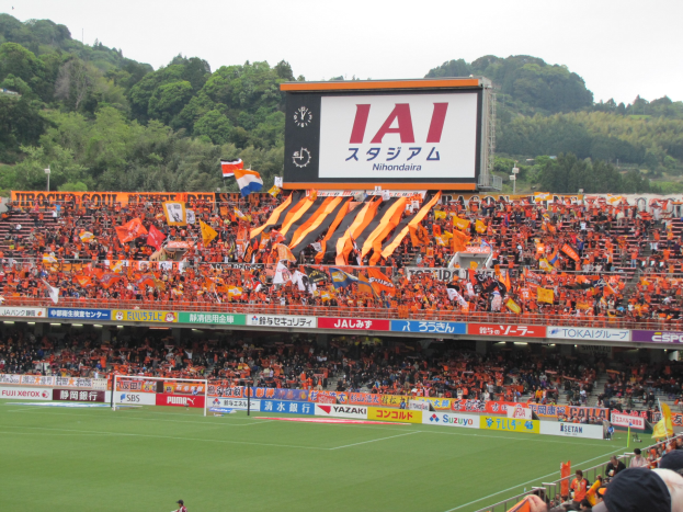 Ein Fußballspiel in einem Stadion mit einer großen Zuschauermenge, grünem Rasen, einem Tor, Bannern, Fahnen, einem großen Bildschirm, Bäumen und einem klaren blauen Himmel.