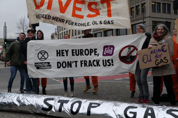 Gruppe von Menschen mit einem "Stoppt Gas"-Schild auf der Straße, die gegen die Europäische Union protestieren, vor dem Hintergrund von Gebäuden, Bäumen, Verkehrsschildern und einem klaren blauen Himmel.