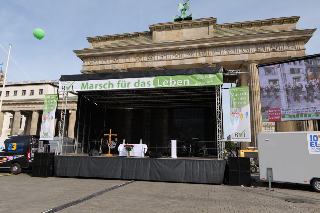 Bühne vor dem Brandenburgertor mit einem Tisch, Bannern, Lautsprechern, Fahrzeugen, Gebäuden, einer Statue, einer Flagge und Wolken.