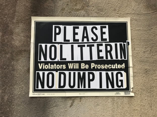 Ein schwarzes und weißes Schild an einem Gebäude trägt die Aufschrift "Bitte Nolitterin' Verursacher werden strafrechtlich verfolgt Kein Entsorgen."