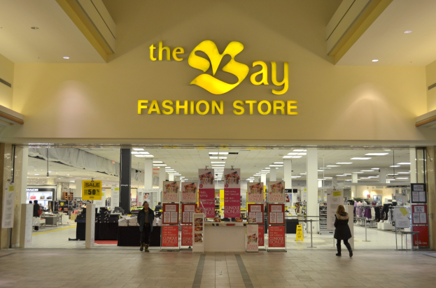 Außenansicht eines Bay Fashion Stores in einem Einkaufszentrum mit Einkäufern, Wandtextanzeigen, Säulen, Deckenleuchten und Hintergrunddekoration.