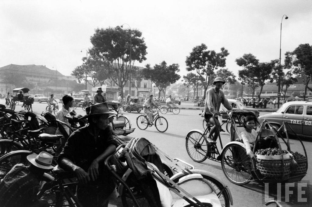 Eine belebte Straße mit Fahrrädern, Autos und anderen Fahrzeugen, die vorbeifahren, mit Bäumen und Gebäuden im Hintergrund.