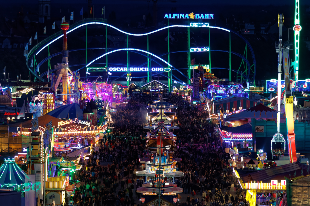 Eine große Menschenmenge versammelte sich um einen hell erleuchteten Karneval in der Nacht, umgeben von Vergnügungsgeräten, Polen, Fahnen und Namensschildern, mit einem dunklen Hintergrund, der eine festliche Atmosphäre schafft.