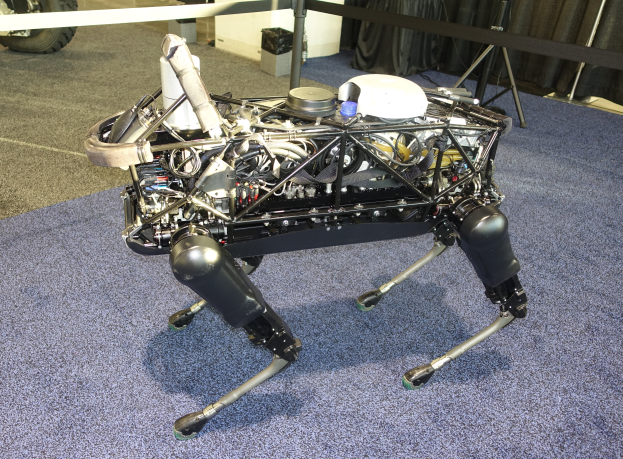 Ein Roboter mit ausgestreckten Armen und Beinen, geneigtem Kopf, sitzt auf einem blauen Teppichboden, mit einer Maschine in der Mitte und einem Fahrzeug auf der linken Seite, umgeben von St├Ąnden, Vorh├Ąngen und anderen Gegenst├Ąnden im Hintergrund.