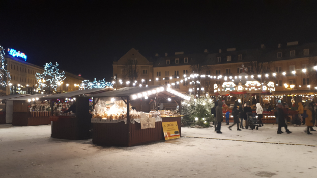 Ein verschneiter nächtlicher Weihnachtsmarkt mit Menschen, Buden, Pflanzen, Bäumen, Gebäuden und Schildern unter einem bewölkten Himmel.